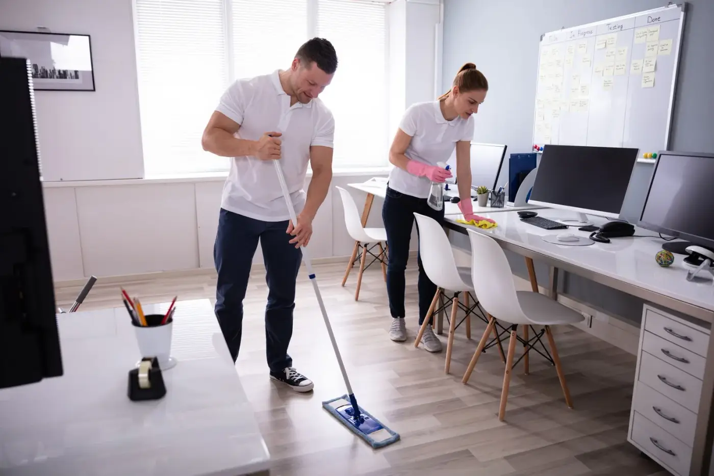 Two people cleaning an office: man mopping floor, woman wiping desk.