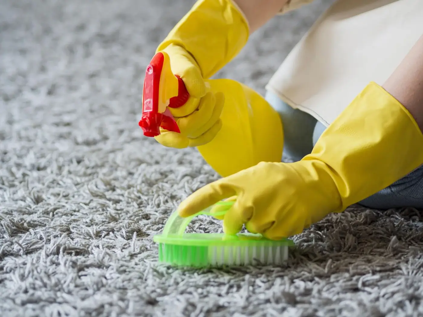 Person wearing yellow gloves cleans a gray carpet with a spray bottle and brush.